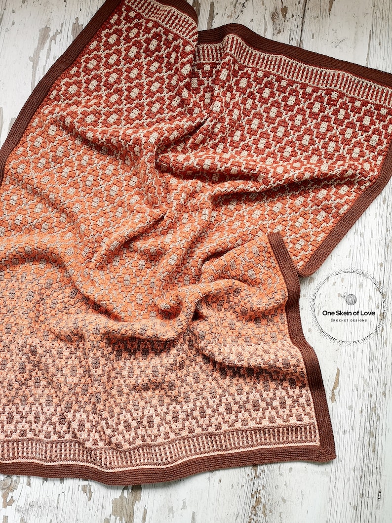 Puede incluir: Una manta de ganchillo con un patr&oacute;n geom&eacute;trico en tonos marrones, naranjas y crema. La manta est&aacute; doblada y drapeada sobre una superficie de madera blanca. El texto "One Skein of Love Crochet Designs" es visible en la esquina inferior derecha de la imagen.