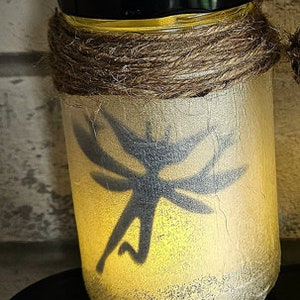 May include: A glass jar with a black lid and a brown twine wrap. The jar is illuminated from within, casting a warm glow. A silhouette of a fairy with wings is visible on the front of the jar.