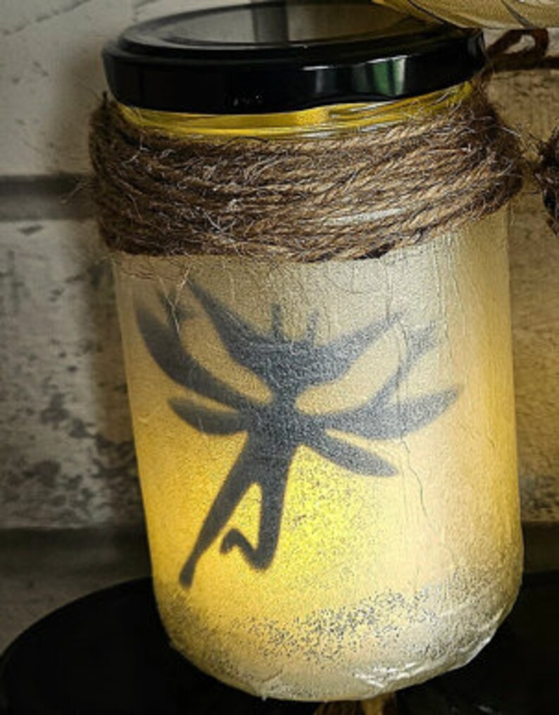 May include: A glass jar with a black lid and a brown twine wrap. The jar is illuminated from within, casting a warm glow. A silhouette of a fairy with wings is visible on the front of the jar.