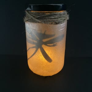 May include: A glass jar with a black lid and a brown twine wrap. The jar is filled with a white substance and has a dragonfly silhouette inside. The jar is lit from within, creating a warm glow.