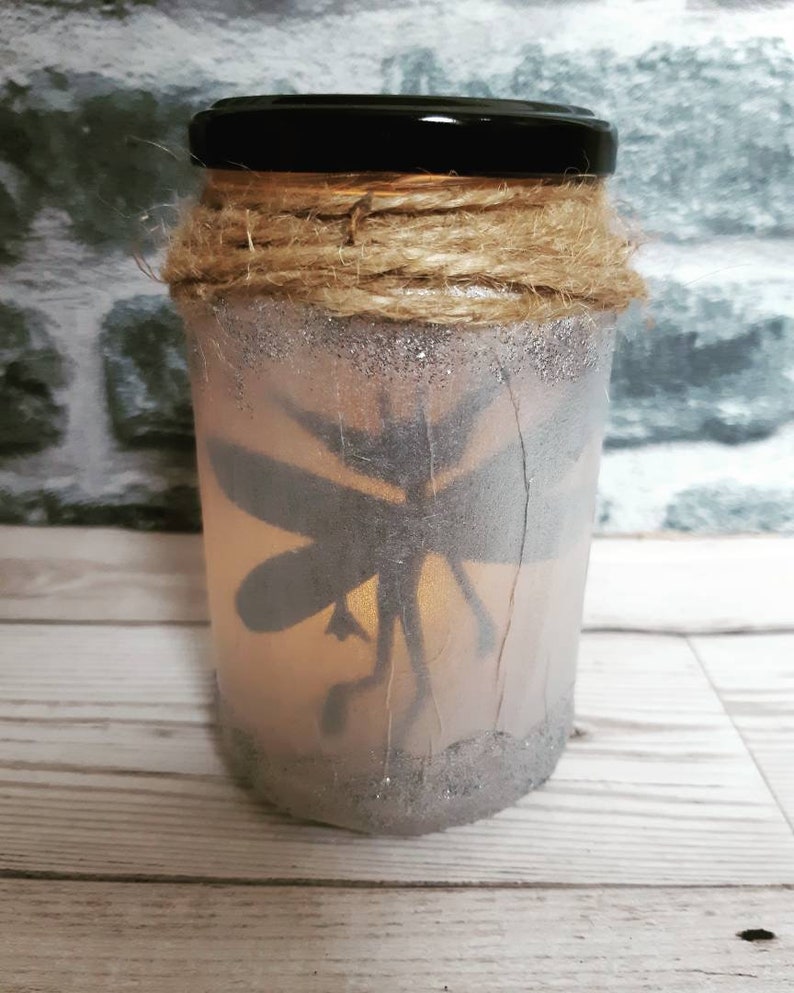 May include: A glass jar with a black lid, decorated with twine and glitter. The jar has a silhouette of a fairy inside, and is lit from within.