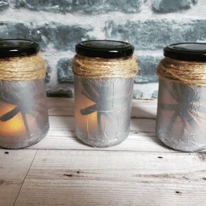 May include: Three silver glass jars with a dragonfly design and a brown twine accent around the top. The jars are lit from inside with a warm white light.