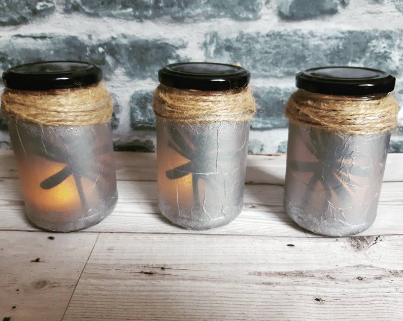 May include: Three silver glass jars with a dragonfly design and a brown twine accent around the top. The jars are lit from inside with a warm white light.