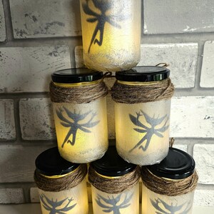 May include: Six glass jars with fairy silhouettes and a warm glow. Each jar is decorated with twine and has a black lid. The jars are stacked in a pyramid shape.