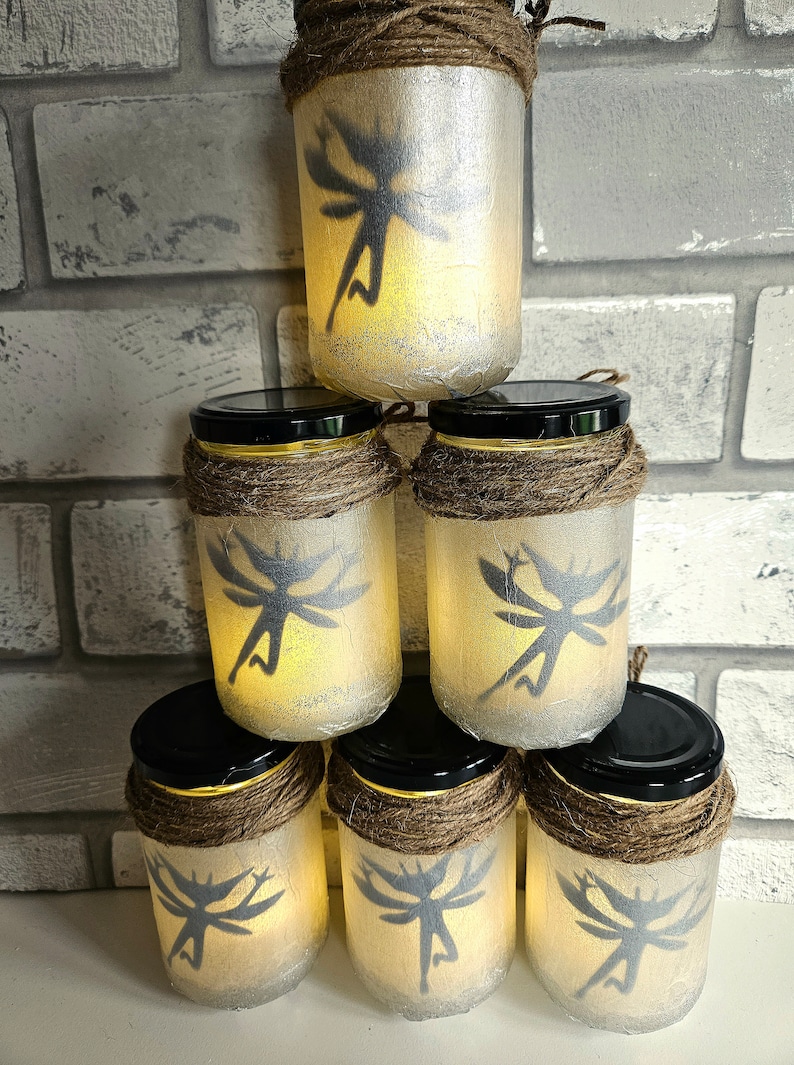 May include: Six glass jars with fairy silhouettes and a warm glow. Each jar is decorated with twine and has a black lid. The jars are stacked in a pyramid shape.