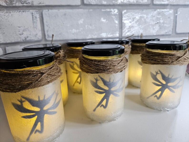 May include: A row of six glass jars with fairy silhouettes, wrapped in twine, and lit from within. The jars are on a white surface.