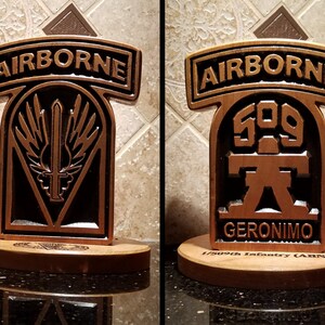 May include: Two wooden plaques with a military theme. The plaque on the left has a black and gold emblem with a sword and wings. The plaque on the right has a black and brown emblem with the number 509 and the word "GERONIMO". Both plaques have the word "AIRBORNE" on top.