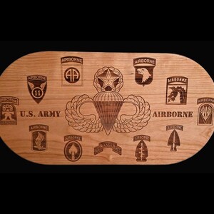 May include: A wooden plaque with various US Army Airborne insignias and the words "U.S. ARMY" and "AIRBORNE" etched into the wood. The plaque is oval-shaped and has a light brown color.