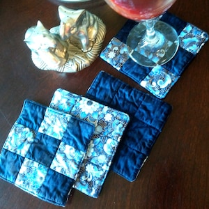 Pode incluir: Quatro  tapetes de mesa acolchoados azul e branco com um padrão floral dourado. Os tapetes de mesa estão dispostos em uma fila sobre uma superfície de madeira.