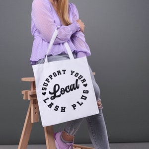 May include: A woman is holding a white tote bag with the text "Support Your Local Plug" printed on it. She is wearing a lavender blouse and light blue jeans.