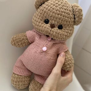 May include: A handmade, plush teddy bear in shades of tan and pink. The bear has a textured body and is wearing a pink knitted outfit with button closures. The bear's eyes are black, and it has a brown nose.