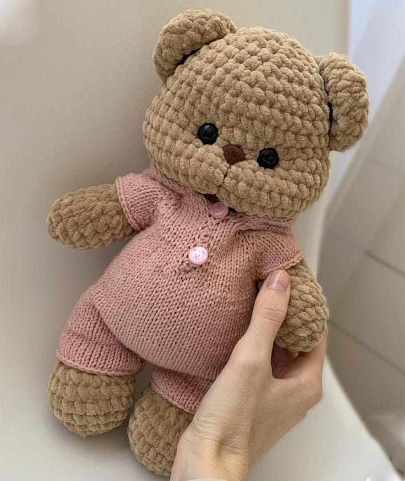 May include: A handmade, plush teddy bear in shades of tan and pink. The bear has a textured body and is wearing a pink knitted outfit with button closures. The bear's eyes are black, and it has a brown nose.