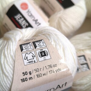 Peut inclure: Pelote de fil blanc avec une &eacute;tiquette indiquant "YarnArt Jeans" et des d&eacute;tails comme le poids (50 g), la longueur (160 m) et la taille des aiguilles (3,5 mm).