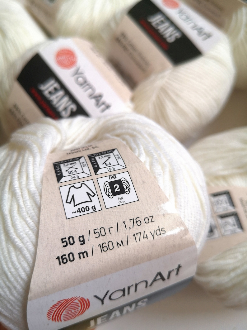 Peut inclure: Pelote de fil blanc avec une &eacute;tiquette indiquant "YarnArt Jeans" et des d&eacute;tails comme le poids (50 g), la longueur (160 m) et la taille des aiguilles (3,5 mm).