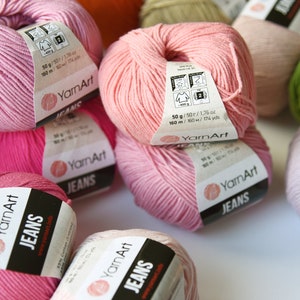 Peut inclure: Gros plan sur plusieurs pelotes de fil rose, vert et violet. Le fil est &eacute;tiquet&eacute; "YarnArt Jeans" et porte les informations de poids et de longueur sur l'&eacute;tiquette. Le fil est compos&eacute; de 50% de coton et de 50% d'acrylique.