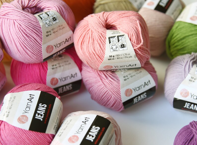 Peut inclure: Gros plan sur plusieurs pelotes de fil rose, vert et violet. Le fil est &eacute;tiquet&eacute; "YarnArt Jeans" et porte les informations de poids et de longueur sur l'&eacute;tiquette. Le fil est compos&eacute; de 50% de coton et de 50% d'acrylique.