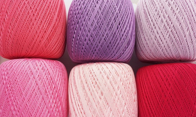 May include: Six balls of yarn in various shades of pink, red, and purple. The yarn is textured and has a delicate pattern.