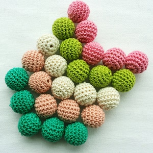 May include: A selection of colourful, crocheted beads in shades of pink, green, cream, and peach. The beads are arranged on a white surface, highlighting their textured, spherical shape. These handmade beads are suitable for jewellery making and craft projects.