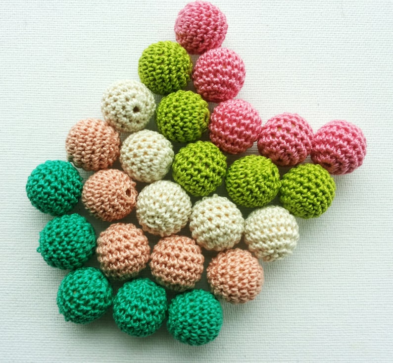 May include: A selection of colourful, crocheted beads in shades of pink, green, cream, and peach. The beads are arranged on a white surface, highlighting their textured, spherical shape. These handmade beads are suitable for jewellery making and craft projects.
