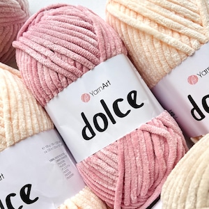 Può includere: Primo piano di diversi gomitoli di filato YarnArt Dolce in vari colori pastello, tra cui rosa e crema. Il filato è morbido e soffice, con una consistenza vellutata. Ogni gomitolo ha un'etichetta bianca con la parola "dolce" in nero.