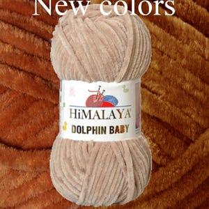 May include: A ball of Himalaya Dolphin Baby yarn in a light brown color. The yarn is soft and plush, with a velvety texture. The label reads "New colors" and features the brand name and product details.