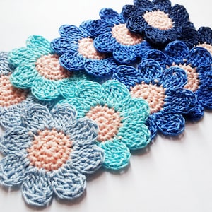 May include: A set of seven crocheted flower appliques in shades of blue and pink. The flowers are arranged in a gradient pattern, with the darkest blue at the top and the lightest blue at the bottom. The flowers have a simple, daisy-like design.