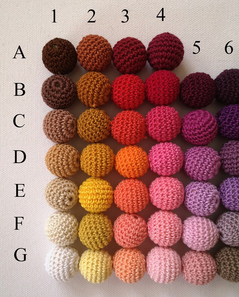 May include: A selection of crochet beads in a grid formation, showcasing a spectrum of colours. The beads vary from dark brown to white, including shades of orange, red, pink, and purple. Each bead is spherical, and the grid is labelled with numbers and letters.