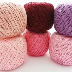 May include: Six balls of yarn in various shades of pink and purple. The balls are stacked in a pyramid shape.