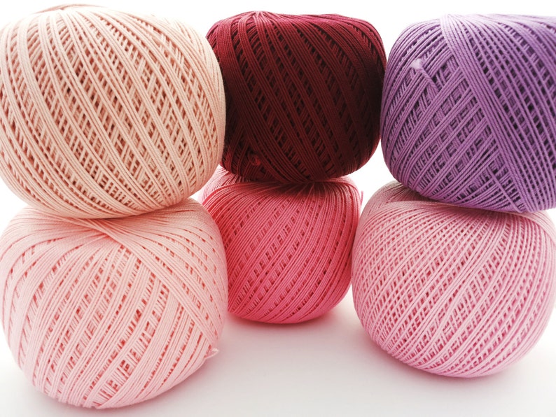 May include: Six balls of yarn in various shades of pink and purple. The balls are stacked in a pyramid shape.