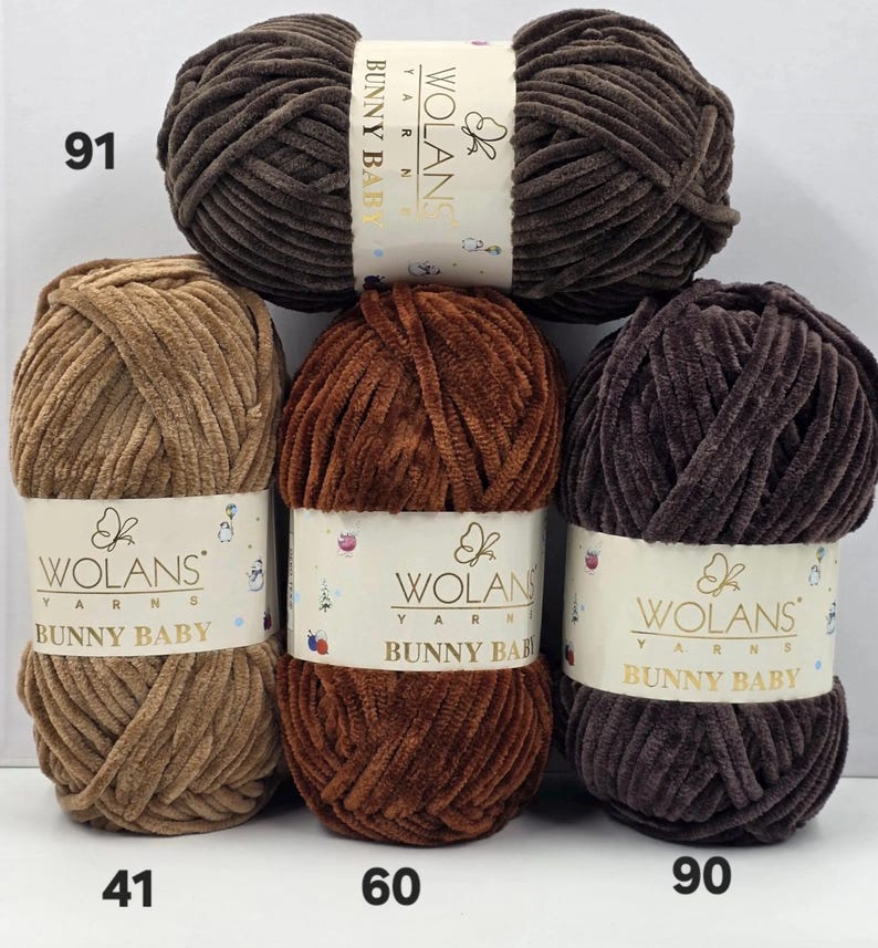 以下が含まれることがあります： ブラウン、タン、ダークチョコレートの色合いのWolans Bunny Baby毛糸玉4個。各毛糸玉には、Wolans Yarnsのブランド名、Bunny Babyの文字、蝶のロゴが印刷された白い紙のラベルが付いています。毛糸は柔らかいベルベットのような質感です。
