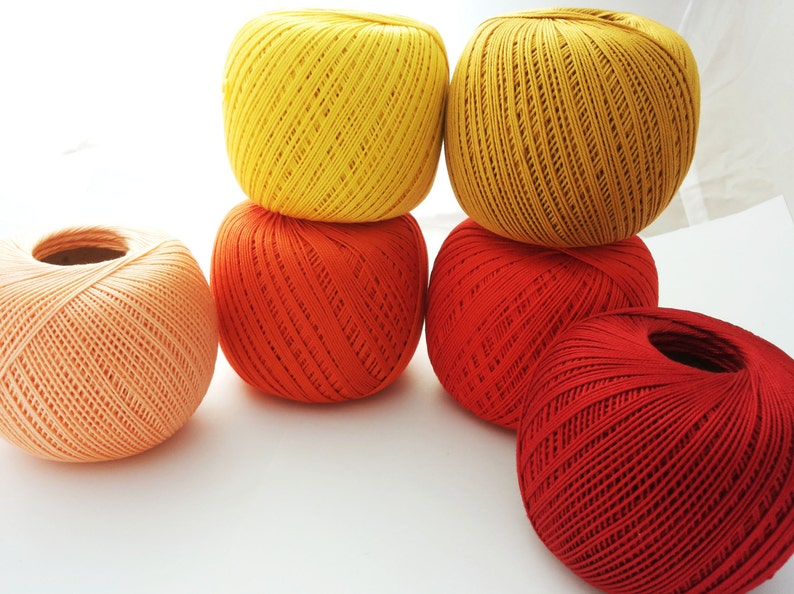 May include: Five balls of yarn in various shades of orange, red, and yellow. The balls are arranged in a pyramid shape.