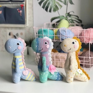 Peut inclure: Trois jouets en crochet en forme de dinosaure. Les dinosaures sont bleu clair, violet et cr&egrave;me avec des accents jaunes, roses et blancs.