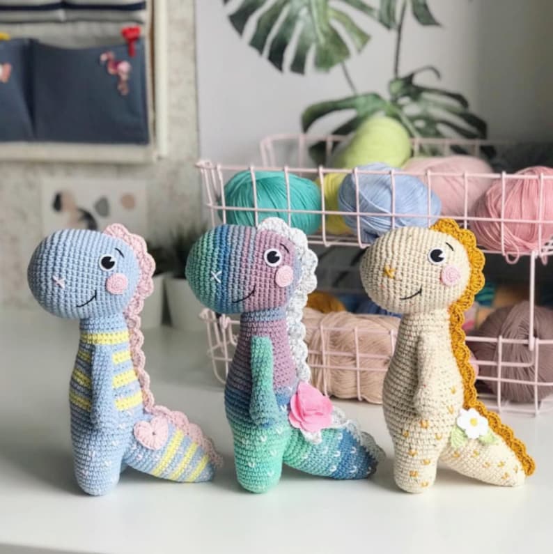 Peut inclure: Trois jouets en crochet en forme de dinosaure. Les dinosaures sont bleu clair, violet et cr&egrave;me avec des accents jaunes, roses et blancs.