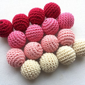 May include: A collection of handmade crochet balls in various shades of pink and cream. The balls are tightly crocheted, creating a textured surface. The colors range from deep red to light pink and cream, perfect for crafting or decorative purposes.