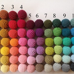 May include: A display of 77 crocheted spheres in a variety of colours, arranged in a grid. Each sphere is labelled with a number and letter for colour identification. The colours span from browns and reds to greens, blues, and purples. The spheres are evenly spaced on a white backdrop.