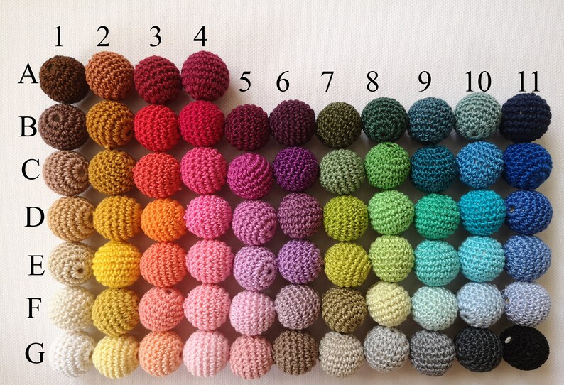 May include: A display of 77 crocheted spheres in a variety of colours, arranged in a grid. Each sphere is labelled with a number and letter for colour identification. The colours span from browns and reds to greens, blues, and purples. The spheres are evenly spaced on a white backdrop.