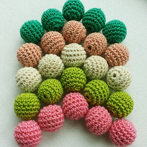 May include: A selection of crochet beads in shades of green, pink, beige, and peach. The beads are closely arranged, highlighting their textured, spherical form. These handmade beads are suitable for craft projects and are ideal for jewellery making.