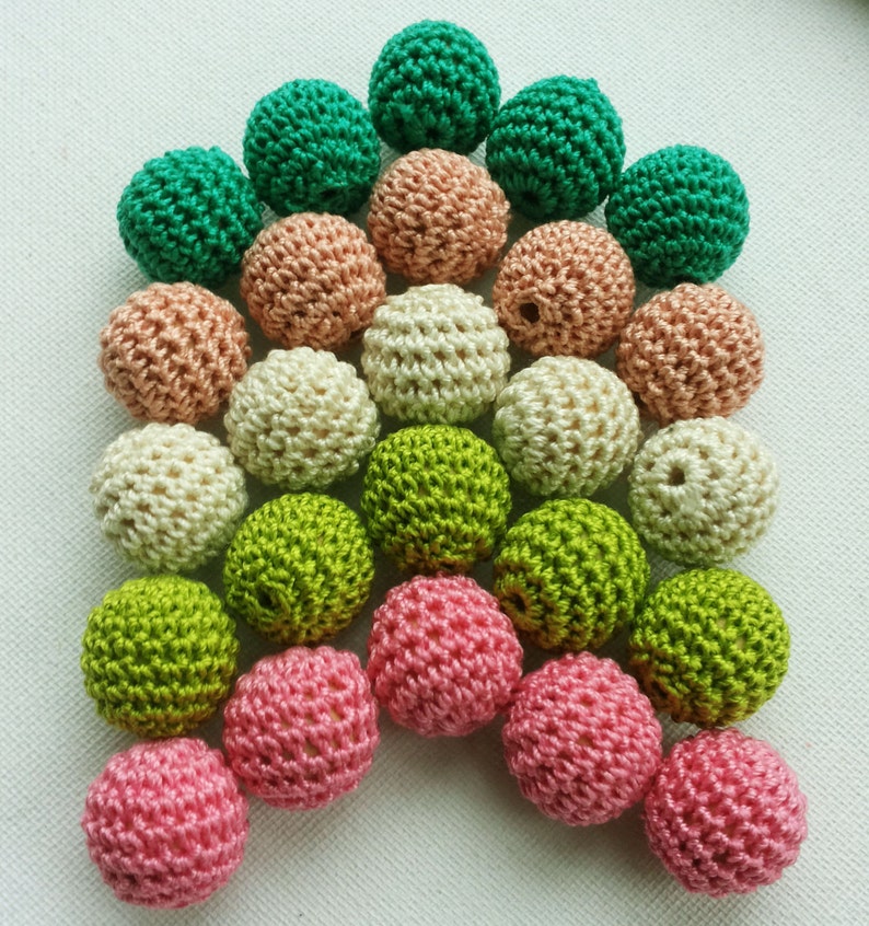 May include: A selection of crochet beads in shades of green, pink, beige, and peach. The beads are closely arranged, highlighting their textured, spherical form. These handmade beads are suitable for craft projects and are ideal for jewellery making.