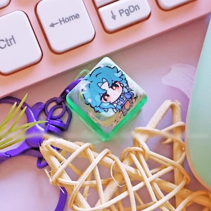 May include: A pastel pink keyboard, a light green game controller, and a decorative star made of woven twigs are arranged on a pink surface. A small, square keychain with a cartoon character is also present, along with a white pen and a purple keychain.