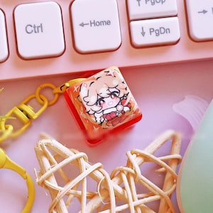 Peut inclure: Un clavier rose pastel, une manette de jeu vert clair et divers objets décoratifs sont disposés sur une surface rose. Un porte-clés jaune, un ornement en forme d'étoile tressée et un stylet blanc sont également présents. Un petit charme avec un personnage d'anime est visible.