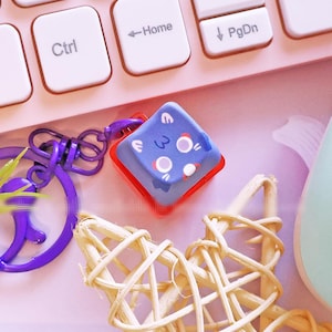 May include: A pastel pink keyboard, a light blue game controller, and a dice-shaped keychain with a purple keyring are arranged on a pink surface. A woven star and a white pen are also present, creating a playful aesthetic.