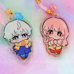 May include: Two anime-style acrylic charms with cute characters. One has white hair and a blue outfit, the other has pink hair and a yellow outfit. Both have gold accents and are on a background of pink and blue feathers.