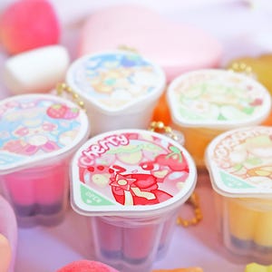 May include: A collection of colorful, small, plastic containers with decorative lids. The lids feature various cartoon designs and the word "Cherry". The containers are filled with different colored jellies.