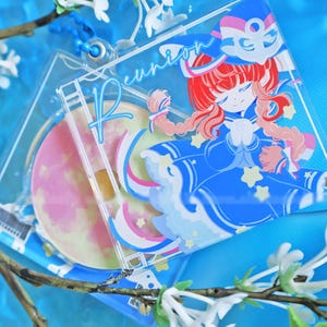 May include: A clear acrylic keyring featuring a cartoon character in a blue outfit with red hair and the word "Reunion". The keyring also includes a circular design with a pink and yellow moon. The background is a light blue.