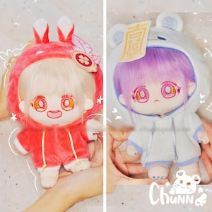 Puede incluir: Se muestran dos muñecos de peluche, uno con un disfraz de conejito rojo y el otro con un disfraz de oso blanco. El muñeco de conejito tiene el pelo rubio y el muñeco de oso tiene el pelo morado. Ambos muñecos tienen ojos grandes y visten trajes con capucha.