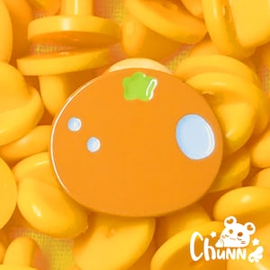 Puede incluir: Pin de esmalte naranja con una hoja verde y dos puntos blancos. El pin tiene un círculo azul claro y está rodeado de objetos amarillos. El pin tiene la palabra "Chunn" en escritura blanca.