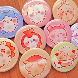 May include: A collection of nine circular pin-back buttons featuring colorful illustrations of various cartoon creatures with expressive faces. The designs include characters in shades of blue, pink, peach, yellow, and green, with one character holding a small white knife.