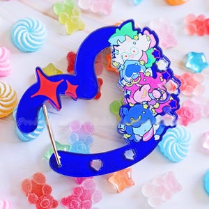 Puede incluir: Un pin azul en forma de corazón con un diseño de personaje de dibujos animados, con un acento de estrella roja. El pin está rodeado de caramelos de colores en varias formas y colores, incluyendo estrellas, osos y espirales.