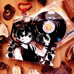 Może przedstawiać: Kwadratowy brelok akrylowy z czarno-białą ilustracją postaci w stylu anime z czerwonymi akcentami liniowymi, zawierający motywy wielu oczu oraz grafikę jajka sadzonego. Brelok posiada czerwoną łańcuszek kulkowy i jest pokazany z etui na płytę CD.