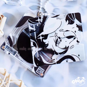 Puede incluir: Dos llaveros de acrílico transparente con ilustraciones en blanco y negro. Uno presenta un personaje estilizado con un sombrero, el otro un diseño de disco de vinilo. Ambos tienen un anillo metálico plateado para sujetar.